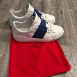 Valentino Garavani vita sneakers - Snygga vita sneakers från Valentino Garavani med bred blå detalj över sidan och snörning. Skorna har en rund tå och platt sula, tillverkade i skinn med diskreta nitar på hälen. Perfekt för dig som gillar exklusiv och stilren design. Boxen är inte i så bra skick.