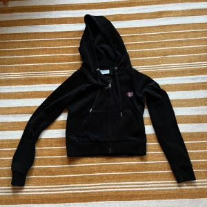 Svart Juicy Couture hoodie i sammet - Svart hoodie från Juicy Couture i mjuk sammet med huva och snörning. Dragkedja framtill, känguruficka och glittrigt hjärta på bröstet. Stor Juicy Couture-logga med krona och hjärta i strass på ryggen. Perfekt för en trendig och chill look.