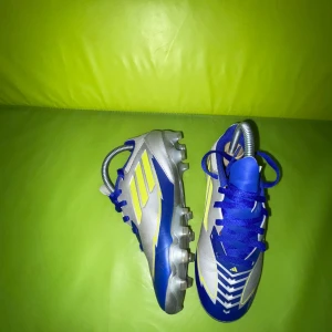 Adidas Messi F50 fotbollsskor blå/gul - Säljer ett par Adidas Messi F50 fotbollsskor i blått, silver och gult med snörning och platt sula med dobbar. Skorna har en sportig design med gula detaljer och Messi-logga på sidan. Perfekta för dig som vill sticka ut på planen.