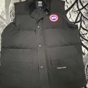 Svart dunväst från Canada Goose - Svart dunväst från Canada Goose med klassisk logga på bröstet och två stora fickor framtill. Västen har hög krage, tryckknappar och är perfekt för lager-på-lager. Snygg och enkel design som funkar till många stilar.