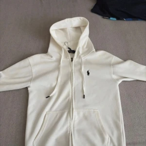 Vit hoodie från Polo Ralph Lauren - Säljer en stilren vit hoodie från Polo Ralph Lauren med dragkedja och klassisk logga på bröstet. Tröjan har huva med snörning och två fickor framtill. Perfekt för en clean och avslappnad look. Använde typ en gång när jag va med tjejen då fick den några makeup fläckar men det går bort med en tvätt