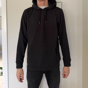 Svart hoodie med huva och ficka - En klassisk svart hoodie i storlek L med huva och snörning. Tröjan har en stor magficka och långa ärmar. Perfekt för en chill och avslappnad stil. Materialet är mjukt och skönt, troligtvis bomull eller polyester.