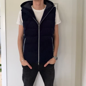 Mörkblå dunväst från Zadolo Stockholm - Snygg mörkblå dunväst från Zadolo Stockholm med huva och hel dragkedja framtill. Västen har ett quiltat mönster och är tillverkad i mjuk manchester på utsidan med slät polyester på insidan. Perfekt för lager-på-lager och streetwear.