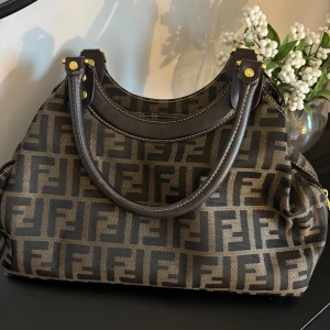 Fendi brun axelväska med FF-mönster - Snygg brun axelväska från Fendi med ikoniskt FF-mönster i svart. Väskan har detaljer i mörkbrunt skinn, guldfärgade metalldetaljer och dragkedja både fram och inuti. Perfekt storlek för att få plats med allt du behöver och en riktig statement piece. Köpt på Vinted 