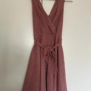 Glittrig rosa omlottklänning Goddiva - Säljer en glittrig rosa kortklänning från Goddiva med omlottdesign och v-ringning. Klänningen har ett matchande bälte i midjan och är ärmlös. Materialet är syntet med glittereffekt och dragkedja bak. Perfekt för dig som vill sticka ut med en snygg look.