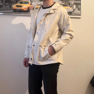 Beige vindjacka från L.L. Bean - Snygg beige vindjacka från L.L. Bean med huva och flera praktiska fickor framtill. Jackan har knappar och dragkedja, samt en clean och stilren look som passar perfekt till vår och höst. Materialet är lätt och känns skönt mot kroppen.