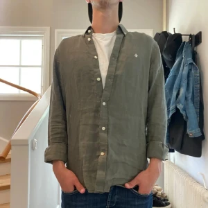 Olivgrön linneskjorta från Morris - Snygg olivgrön skjorta från Morris i ren linne, perfekt för dig som gillar en chill och stilren look. Skjortan har klassisk krage, vita knappar och lång ärm. Diskret Morris-logga broderad på bröstet. Passar dig som vill ha en luftig och bekväm skjorta.