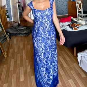 Blå långklänning med blommigt mönster - Säljer en elegant långklänning i blått med blommigt spetsmönster och fyrkantig ringning. Klänningen har breda axelband och är figurnära.