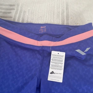 Blå och rosa träningsleggings Crivit M - Snygga blå träningsleggings från Crivit i storlek M med diskret geometriskt mönster och rosa midjeband. Byxorna är trekvartsmodell och har en tight passform, perfekta för gymmet eller löprundan. Materialet är stretchigt och andas, vilket ger skön rörelsefrihet.