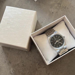 Poedagar silverfärgad klocka - Stilren klocka från Poedagar med svart urtavla och dag/datumvisning. Klockan har ett klassiskt runt boett i silverfärgat metall och matchande länkarmband. Quartz-urverk och minimalistisk design gör den enkel att bära varje dag.