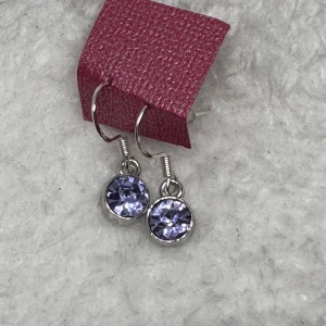 Silverörhängen med lila stenar - Snygga örhängen i silverfärgad metall med runda, facetterade lila stenar som glittrar. Perfekt för dig som vill ha en enkel men ändå iögonfallande detalj till din look. Hängande modell med klassisk krok.
