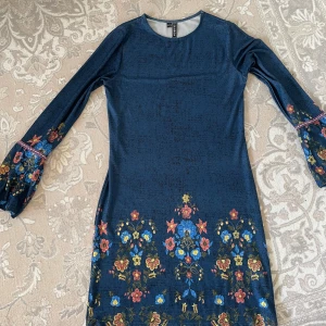 Blommig blå klänning från Rainbow - Supersöt blå klänning från Rainbow med färgglada blommor i gult, rött och blått längs nederkant och ärmar. Långärmad med lätt utsvängda ärmslut och dekorativt band. Mjuk, stretchig polyester som sitter bekvämt och har en normal passform.