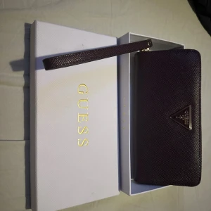 Mörklila plånbok från Guess - Snygg plånbok från Guess i mörklila med struktur och metalllogga framtill. Plånboken har ett handledsband och dragkedja runt om, perfekt för att hålla koll på dina grejer. Kommer i originalask med Guess-logga i guld.