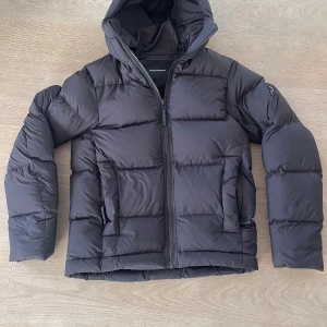 Svart pufferjacka Peak Performance - Säljer en svart pufferjacka från Peak Performance i storlek S. Jackan har huva, dragkedja framtill och två sidofickor med dragkedja. Den är quiltad och har en clean, modern look som passar perfekt när det är kallt ute. Jackan är köpt från förra året och är i ny skick. Ordinarie pris 4200