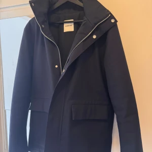 Marinblå vinterjacka från Sandro med huva - Stilren svart vindjacka från Sandro med huva, dragkedja och tryckknappar framtill. Jackan har två stora fickor med lock och en minimalistisk design. Perfekt för dig som gillar en clean look och vill ha något som funkar till många outfits. Den är i storlek S och kan även passa dig med M. Ny pris är 4999kr 