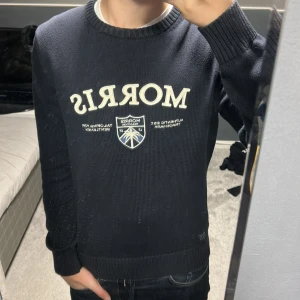 Mörkblå stickad tröja från Morris - Säljer en mörkblå stickad tröja från Morris med vit broderad logga och emblem på bröstet. Tröjan har rund halsringning med ribbade muddar och en snygg detalj med randig insida i halsen. Perfekt för dig som gillar klassisk och stilren look.