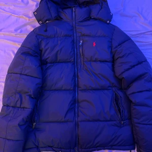 Blå pufferjacka från Polo Ralph Lauren - Säljer en blå pufferjacka från Polo Ralph Lauren med huva och dragkedja framtill. Jackan har en röd Polo-logga på bröstet, flera fickor med dragkedja och quiltad design. Perfekt för kalla dagar och har en normal passform. 