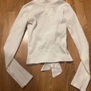Vit ribbad topp med knyt från Ginatricot - Superfin vit topp från Ginatricot i ribbad bomull med långa ärmar och två snygga knytband baktill. Toppen har en v-ringad design och croppad passform som ger en trendig look. Perfekt för dig som gillar stilrena detaljer och vill sticka ut.