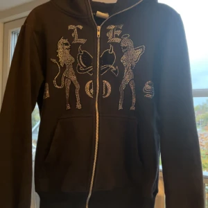 Svart hoodie med strass från Le Dos - Svart hoodie från Le Dos med dragkedja och huva. Framsidan är dekorerad med stora strassmotiv av två figurer och text. Tröjan har långa ärmar och känguruficka framtill. Perfekt för dig som gillar statement-plagg med bling.