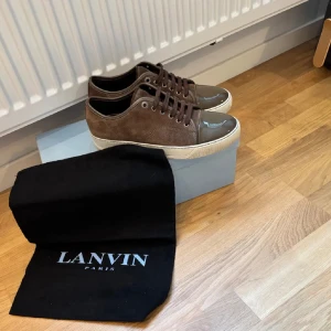 Bruna sneakers från Lanvin Paris - Snygga bruna sneakers från Lanvin med ovandel i mocka och lackad tå i mörkbrunt. Skorna har vita gummisulor och bruna snören. Klassisk låg modell med rund tå och diskreta metalldetaljer vid snörningen. Perfekta för dig som gillar stilrena och lyxiga sneakers.box en dustbag och nya snören till dom skriv vid minst fundering. Nästan oanvänd 
