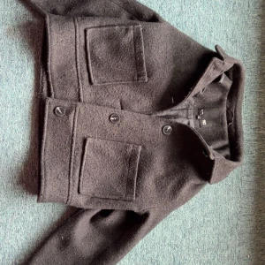 Svart ullkappa från 157, XS - Kort svart kappa från 157 i ull med två stora fickor framtill och knäppning med knappar. Klassisk krage och stilren design som passar perfekt till höst och vinter. Snygg och enkel att matcha med olika outfits.