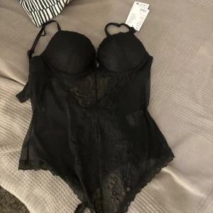 Svart spetskorsett med push up bh - Säljer en svart korsett med vadderade kupor, spetsdetaljer och fina rosetter på sidorna. Axelbanden är justerbara och materialet är en mix av spets och mesh för en snygg look. Perfekt för dig som vill ha något extra till outfiten.