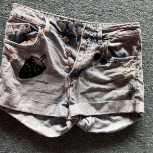 Ljusblå jeansshorts med jordgubbstryck - Säljer ett par ljusblå jeansshorts från H&M Divided med coola svarta jordgubbstryck och slitna detaljer. Shortsen har knäppning med flera knappar framtill, uppvikta benslut och klassiska fickor. Perfekta för sommaren!