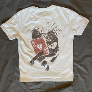 Vit t-shirt från Masteme - Vit t-shirt från Masteme med coolt tryck på ryggen av ett ansikte som håller ett rött hjärtkort och texten 'Masteme' i svart på bröstet. Klassisk rund hals och korta ärmar. Materialet känns som mjuk bomull, perfekt för en chill och edgy stil. Nypris 600kr knappt använd