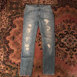 Ljusblå slitna jeans från Lee 30/32 - Ljusblå jeans från Lee med snygga slitningar och hål på framsidan. Klassisk femficksmodell med raka ben och normal passform. Jeansen har Lee-logga på bakfickan och är tillverkade i mjukt denimtyg. Perfekta för en avslappnad streetstil. Midja 37cm längd 100cm (63)
