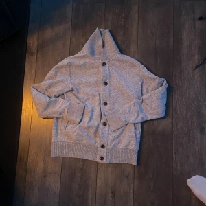 Grå kofta med hög krage och knappar - Snygg gråmelerad kofta med hög ribbad krage och stora knappar framtill. Tröjan har långa ärmar och breda muddar vid ärmslut och nederkant. Perfekt för lager-på-lager och chill dagar.