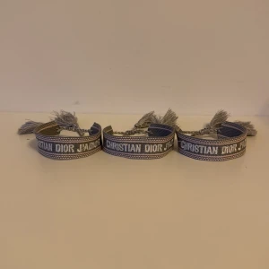 Grå Christian Dior armband med tofsar - Säljer tre snygga armband från Christian Dior i grått och vitt med broderad logga och coola tofsar i ändarna. Armbanden har en bred, vävd design som ger en trendig och exklusiv känsla. Perfekt accessoar för att lyfta din outfit.125kr styck 300kr för alla