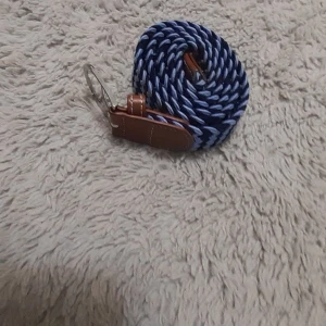 Randigt blått och vitt bälte - Säljer ett snyggt randigt bälte i blått och vitt med bruna skinn-detaljer och silverfärgat spänne. Bältet har en flätad design som ger en cool och modern look. Perfekt accessoar för att lyfta din outfit.