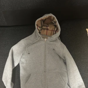 Grå hoodie från Burberry med dragkedja - Snygg grå hoodie från Burberry med klassiskt rutigt foder i huvan. Tröjan har dragkedja framtill, fickor och justerbar huva med snörning. Mjuk insida och stilren look, perfekt för dig som gillar exklusiva streetwear-vibbar.