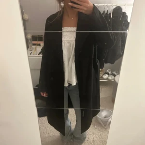 Svart lång kappa - Säljer en svart, stilren och lång kappa med bälte i midjan. Jackan har en klassisk rak passform och är perfekt för dig som gillar en enkel men snygg look. Passar till jeans och har långa ärmar. Fråga gärna om fler bilder eller mått!