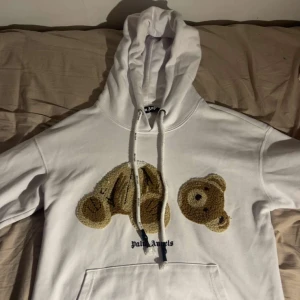 Vit Palm Angels hoodie med björn - Skaffa dig en vit hoodie från Palm Angels med ikoniskt björnmotiv framtill. Tröjan har huva med snörning, känguruficka och broderad logga. Tillverkad i polyesterblandning, perfekt för dig som vill sticka ut med streetwear-vibe.