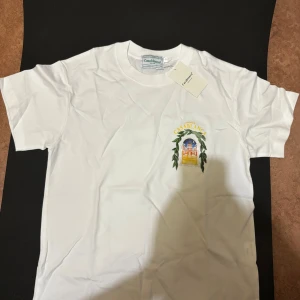 T-shirt från Casablanca med tryck - Vit t-shirt från Casablanca med färgglatt tryck av en byggnad och gröna blad både fram och bak. Klassisk rund hals och korta ärmar. Mjuk bomullskvalitet och snygg streetstil. Perfekt för dig som gillar unika prints och vill sticka ut.