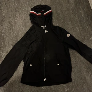 Svart vindjacka från Moncler med huva - Svart vindjacka från Moncler med huva och dragkedja framtill. Jackan har Moncler-logga på ärmen och snygga rödvita detaljer på huvan. Två fickor med tryckknappar och stilren design. Perfekt för dig som vill ha en clean och sportig look. STOCKHOLM bara!! STORLEK S