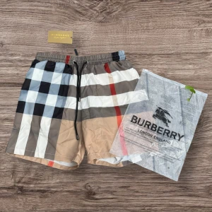 Burberry rutig badshorts med snörning - Snygga badshorts från Burberry med klassiskt rutigt mönster i beige, svart, vit, blå och röd. Shortsen har elastisk midja med svart snörning och meshfoder på insidan. Perfekta för sommaren och strandhäng. En bakficka ger extra stilpoäng.