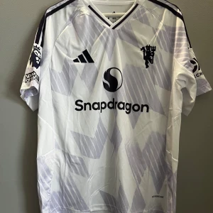 Vit Manchester United Cunha 10 tröja - Säljer en vit Manchester United fotbollströja från Adidas med lila geometriskt mönster, svarta detaljer och tryck. Tröjan har korta ärmar, V-ringning och sponsorloggor som Snapdragon. Namn och nummer 'Cunha 10' på ryggen. Tillverkad i lätt funktionsmaterial. Kan lösa fler bilder 