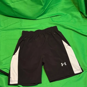 Svarta träningsshorts från Under Armour - Svarta träningsshorts från Under Armour med vita sidopaneler och elastisk midja med snörning. Perfekta för gymmet eller löprundan, tillverkade i lätt och snabbtorkande syntetmaterial. Klassisk logga på vänster ben och normal passform.Liknande modell som originalet!