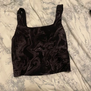 Svart och lila mönstrad linnetopp - Cool linnetopp med svart bas och lila marmorerat mönster. Toppen har breda axelband och croppad passform, perfekt till jeans eller kjol. Materialet känns mjukt och stretchigt, vilket gör den bekväm att bära hela dagen.