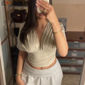 Beige croppad topp med v-ringning - Säljer en beige croppad topp med bred v-ringning. Toppen har korta, vida ärmar och snygga rynkningar vid midjan som ger en cool siluett. Perfekt för dig som gillar trendiga och bekväma plagg med lite extra edge.