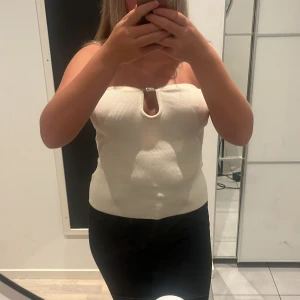 Vit ribbad tubtopp från H&M - Säljer en vit ribbad tubtopp från H&M med en snygg cut-out och metallring framtill. Toppen är axelbandslös och har en tight passform som framhäver figuren. Perfekt för dig som gillar stilrena och trendiga plagg.