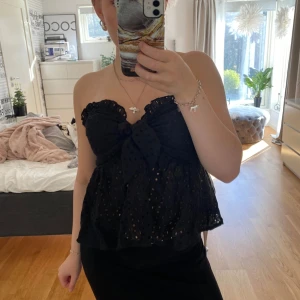 Svart bandeau tubtopp med rosett - Supersöt svart bandeau tubtopp med broderade hål och stor rosett framtill. Toppen är axelbandslös och har volangkant upptill och nertill för en extra feminin vibe. Perfekt för dig som vill sticka ut med en unik och trendig topp.