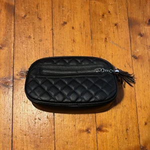 Svart axelväska med mönstrad rem - Snygg svart axelväska i skinnimitation. Den har två fack i sig. Sen finns de ett fack på utsidan. Perfekt för dom som inte har med sig mycket så passar den dig. 