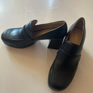 Svarta skinnloafers med klack från Other Stories - Snygga svarta loafers från & Other Stories i en klassisk design med rund tå och bred rem över foten. Tillverkade i skinn som ger en stilren look. Perfekta för dig som gillar retrovibbar och vill ha lite höjd.