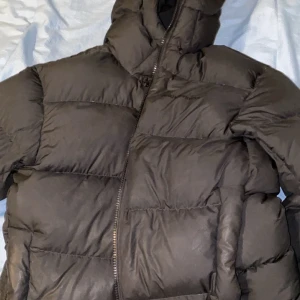 Svart pufferjacka från Peak Performance - Säljer en svart pufferjacka från Peak Performance med hög krage och huva. Jackan har breda, quiltade sektioner och dragkedja framtill. Perfekt för kalla dagar och har en clean, modern look som funkar till de flesta stilar.