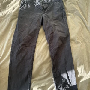 Svarta chinos från Dressmann slim fit - Svarta chinos från Dressmann i slim fit-modell. Byxorna har klassisk knapp och dragkedja framtill, samt snedställda fickor på sidorna. Materialet är mjukt och bekvämt, perfekt för en clean och stilren look.