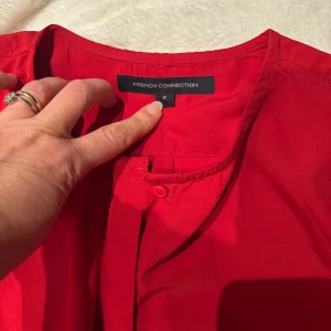 Röd henley från French Connection - Snygg röd henley-skjorta från French Connection i storlek M. Skjortan har lång ärm, rund hals och knappar framtill. Volang fram på ena sidan. Tillverkad i mjuk återvunnen polyester som känns skön mot huden. Perfekt för dig som gillar färgstarka plagg med enkel design.