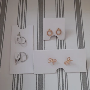 Tre par örhängen i silver och guld - Säljer tre par örhängen: ett par små silvriga hoops med enkel design, ett par guldiga cirklar med tvinnad struktur och ett par guldiga rosettformade studs. Perfekt för dig som gillar att mixa och matcha olika stilar och färger.